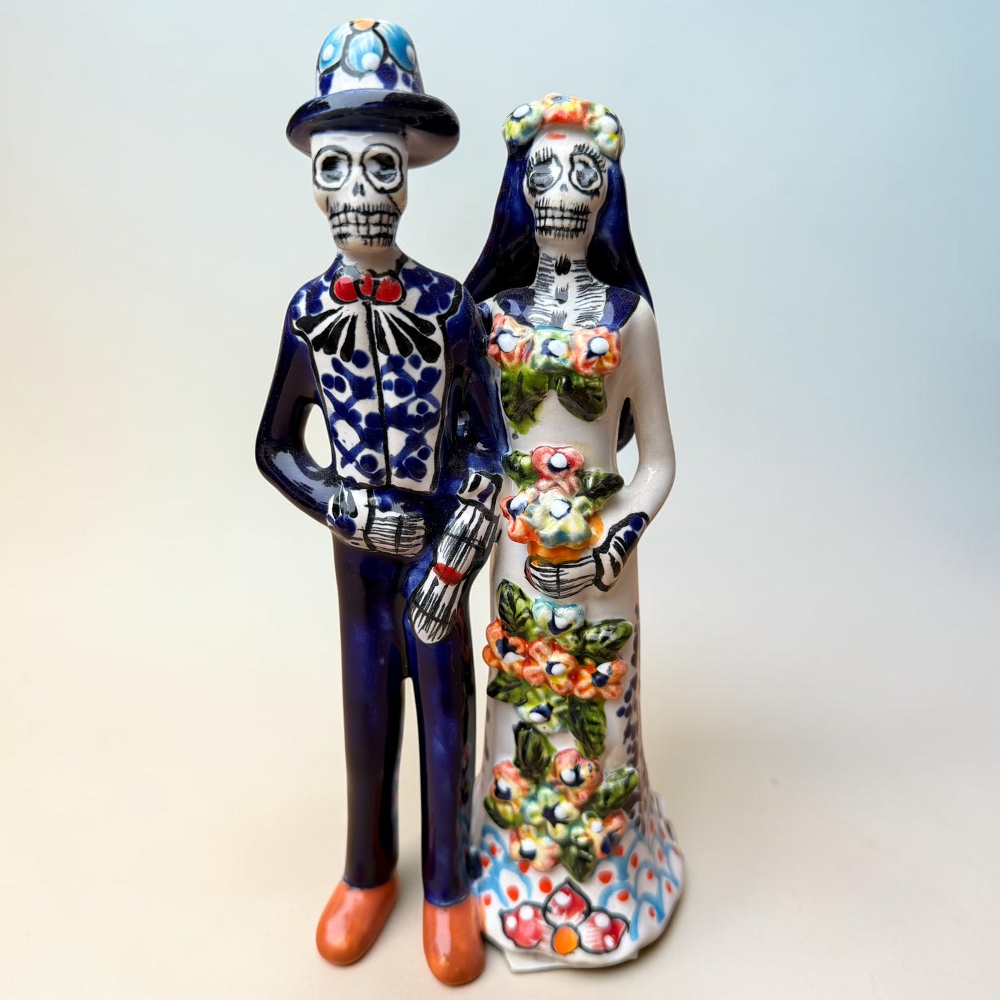A hand-painted Talavera Day of the Dead bride and groom figurine. The groom is dressed in a blue suit with white patterns, a black bow tie, and an orange-red bow tie accent, wearing a blue hat. The bride is dressed in a dress adorned with flowers and wearing a veil.