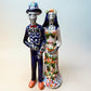 A hand-painted Talavera Day of the Dead bride and groom figurine. The groom is dressed in a blue suit with white patterns, a black bow tie, and an orange-red bow tie accent, wearing a blue hat. The bride is dressed in a dress adorned with flowers and wearing a veil.