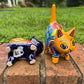 Two colorful talavera ceramic cat figurines standing on a brick surface outdoors with grass and trees blurred in the background. One cat is dark blue with orange, yellow, and red patterns, and the other is orange with blue, red, yellow, and white patterns. This is the bottom view.