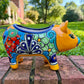 A colorful ceramic pig planter with a yellow-orange face and body, decorated with blue, green, red, and white floral patterns, sitting on a brick surface with a blurred outdoor garden background. View of left hand side of pig.