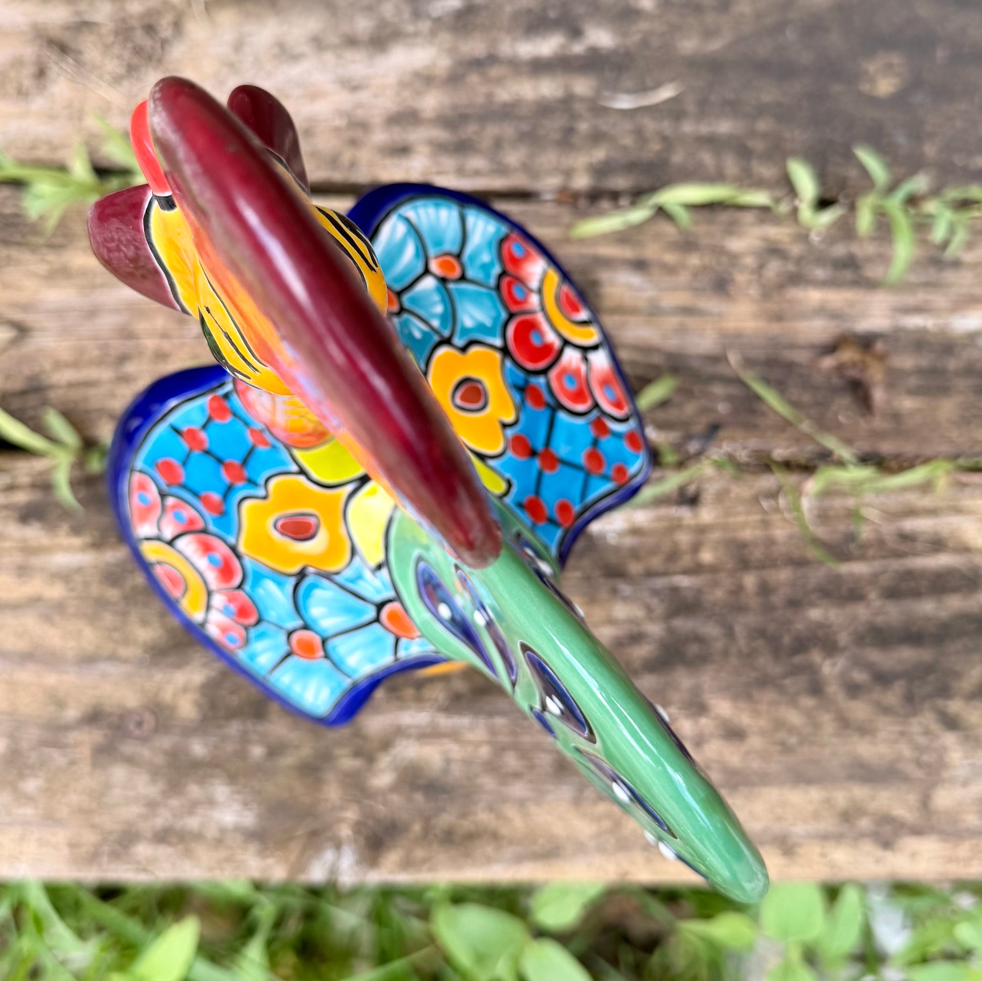 A colorful hand-painted ceramic Mexican-style rooster figurine standing on a rustic wooden surface outdoors, top of rooster view. 