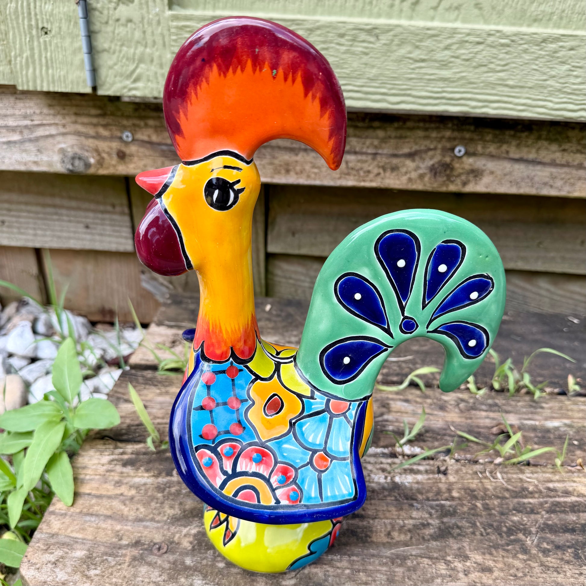 A colorful hand-painted ceramic Mexican-style rooster figurine standing on a rustic wooden surface outdoors, right side view.