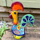 A colorful hand-painted ceramic Mexican-style rooster figurine standing on a rustic wooden surface outdoors, right side view.