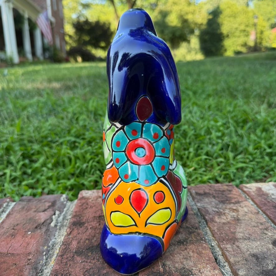 A colorful hand-painted talavera ceramic coyote figurine sitting on a brick surface with a blurred green lawn and trees in the background back view