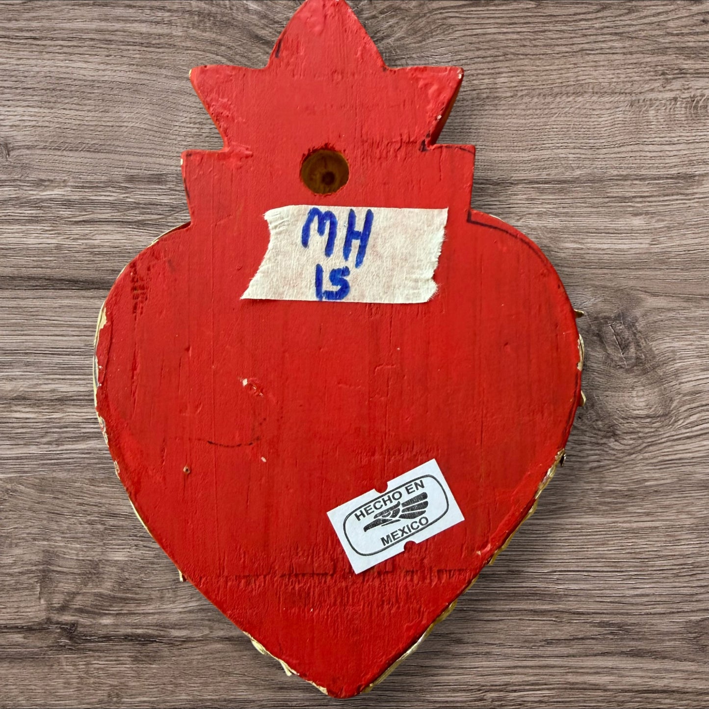 A Milagros heart featuring a gold textured top and a red heart base covered in raised silver-toned charms displayed on a wooden wall, back view.