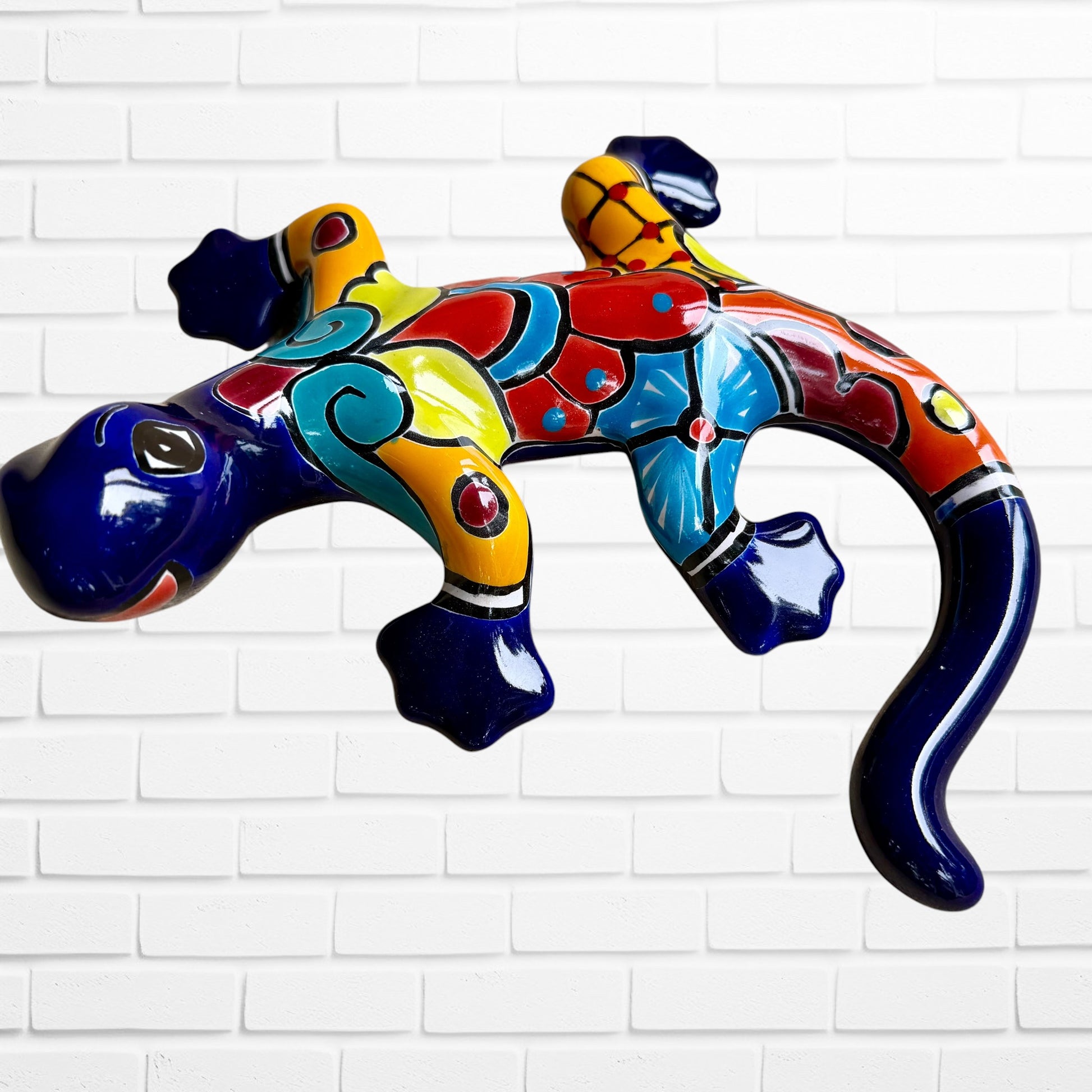 A colorful ceramic wall sculpture of a stylized lizard or salamander, decorated with vibrant hand-painted patterns in blue, red, yellow, and orange, mounted on a white brick wall front face.
