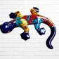 A colorful ceramic wall sculpture of a stylized lizard or salamander, decorated with vibrant hand-painted patterns in blue, red, yellow, and orange, mounted on a white brick wall front face.
