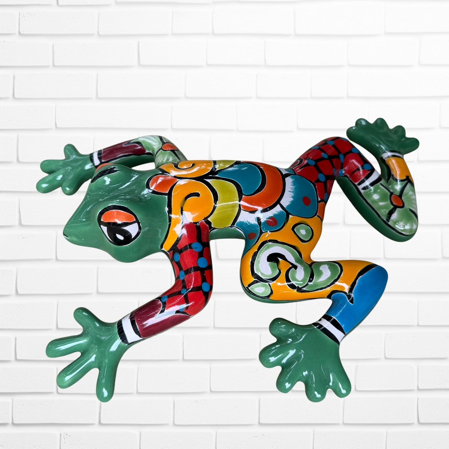 A colorful hand-painted ceramic frog wall sculpture in a dynamic jumping pose, decorated with vibrant green, red, orange, yellow, and blue patterns, set against a white brick wall background side view.