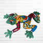 A colorful hand-painted ceramic frog wall sculpture in a dynamic jumping pose, decorated with vibrant green, red, orange, yellow, and blue patterns, set against a white brick wall background side view.