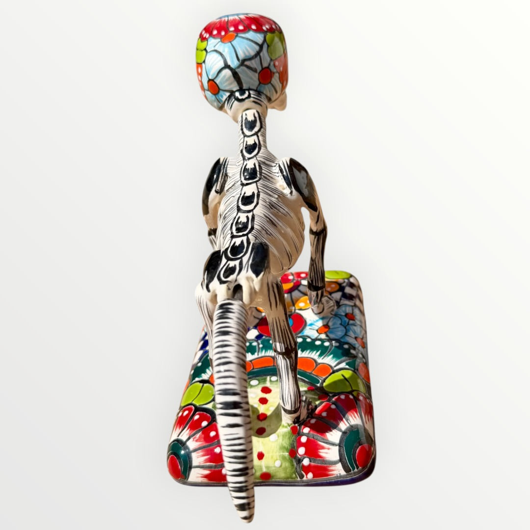 A ceramic Mexican-style skeleton dog figurine standing on a colorful floral base against a plain white background. Back view of catrina skeleton dog showing backbone