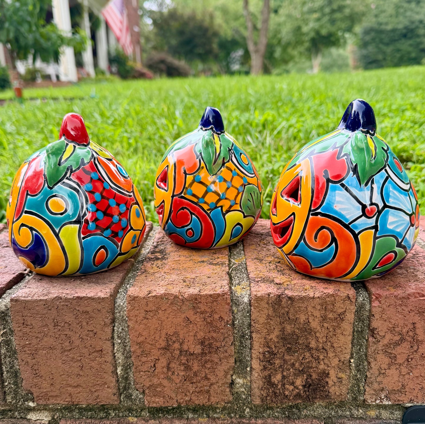 Three hand-painted Talavera ceramic jack-o'-lantern pumpkins in bright orange, yellow, red, green, and blue, sitting on a brick ledge with a blurred green lawn and house in the background, side view