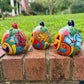 Three hand-painted Talavera ceramic jack-o'-lantern pumpkins in bright orange, yellow, red, green, and blue, sitting on a brick ledge with a blurred green lawn and house in the background, side view