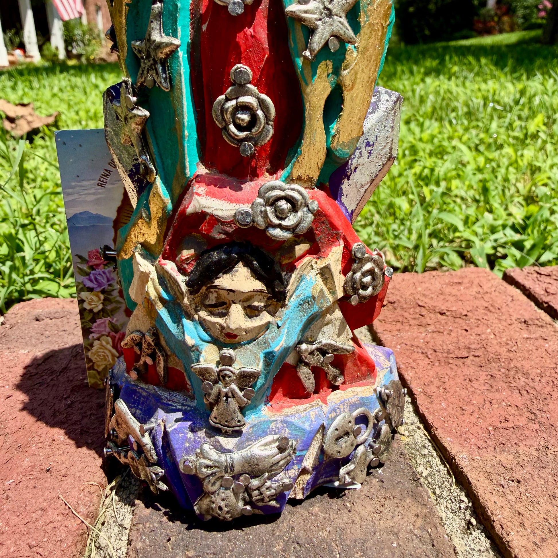 A colorful Milagros Guadalupe  (Virgin Mary) standing on a brick surface outdoors with a grassy yard in the background, close-up of bottom child.