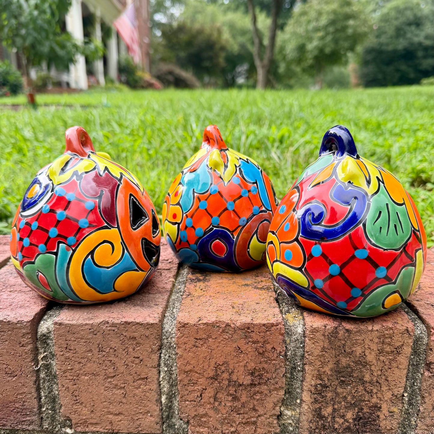 Three hand-painted Talavera ceramic jack-o'-lantern pumpkins with orange faces and yellow, blue, and red green designs, sitting on a brick path in a grassy backyard, left side view.