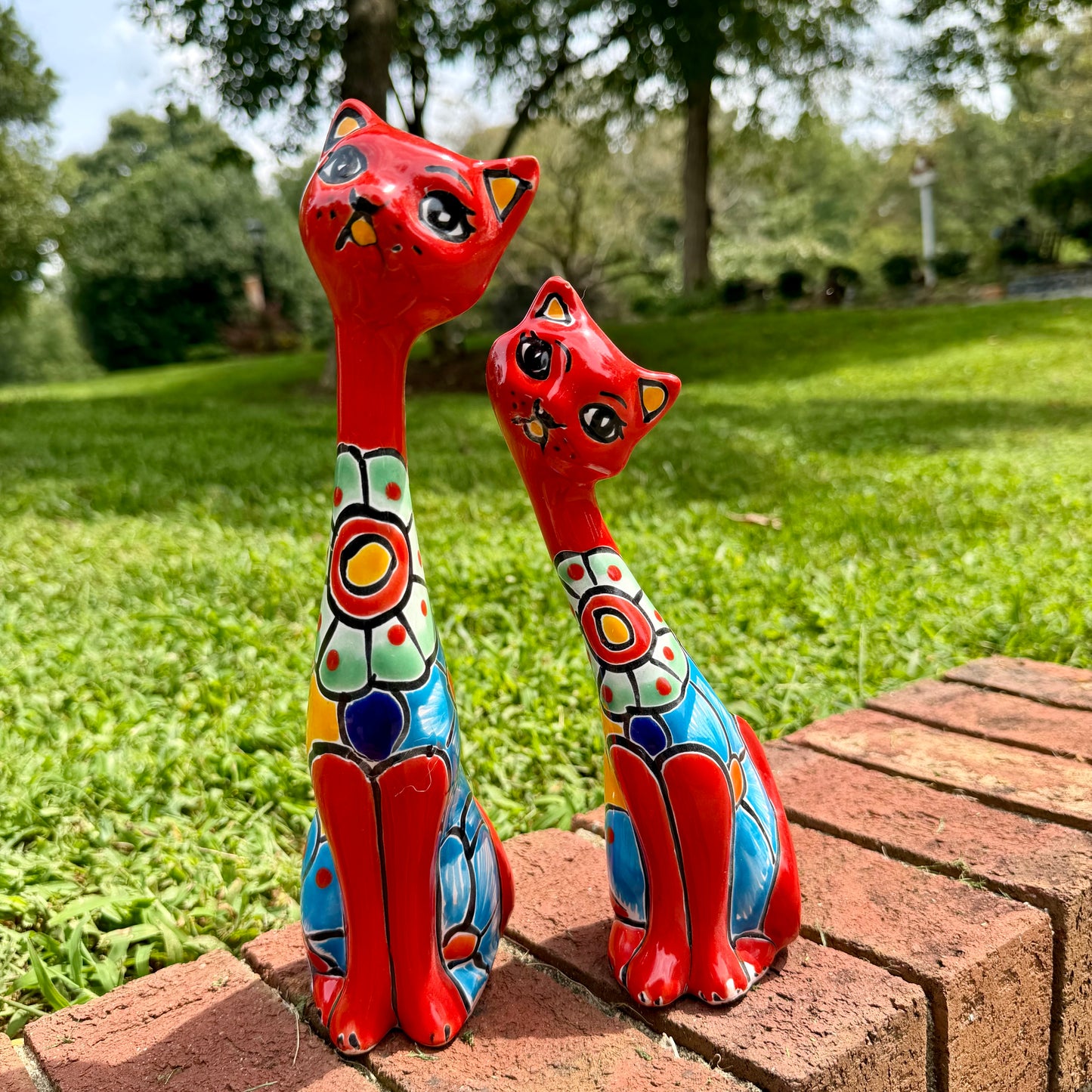 Two hand-painted ceramic cat figurines in red and blue with colorful geometric patterns, sitting on a brick path in a grassy park.