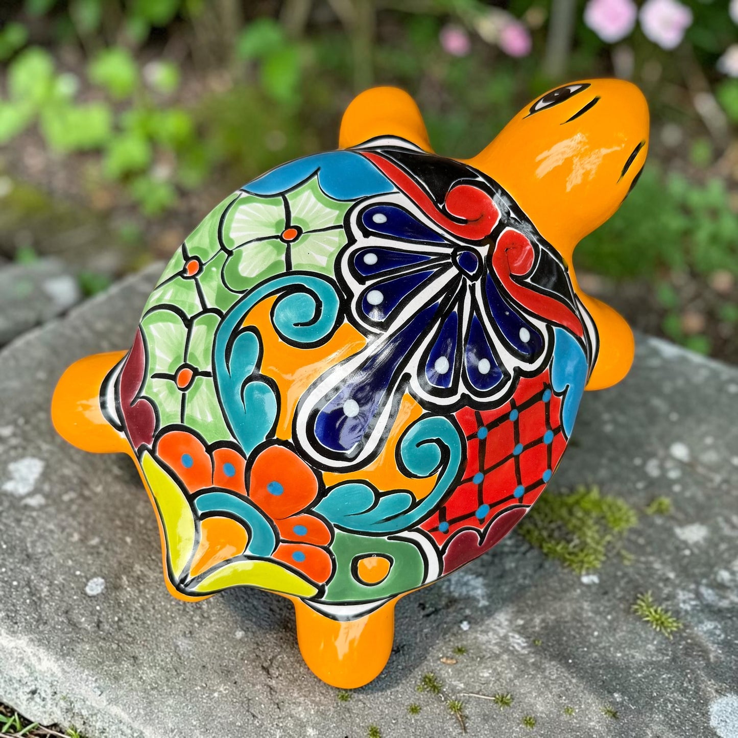 A glossy ceramic turtle figurine with a bright yellow body and a highly decorated shell in red, blue, yellow, black, and white, resting on a stone surface with a blurred garden background, top shell view.