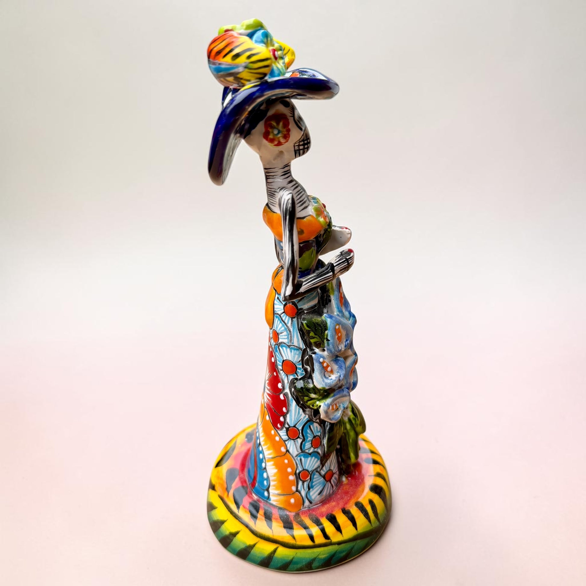 A Talavera Day of the Dead (Día de los Muertos) ceramic skeleton figurine with calla Lillies standing on a patterned base against a soft pink gradient background. This is her left side view.