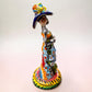 A Talavera Day of the Dead (Día de los Muertos) ceramic skeleton figurine with calla Lillies standing on a patterned base against a soft pink gradient background. This is her left side view.