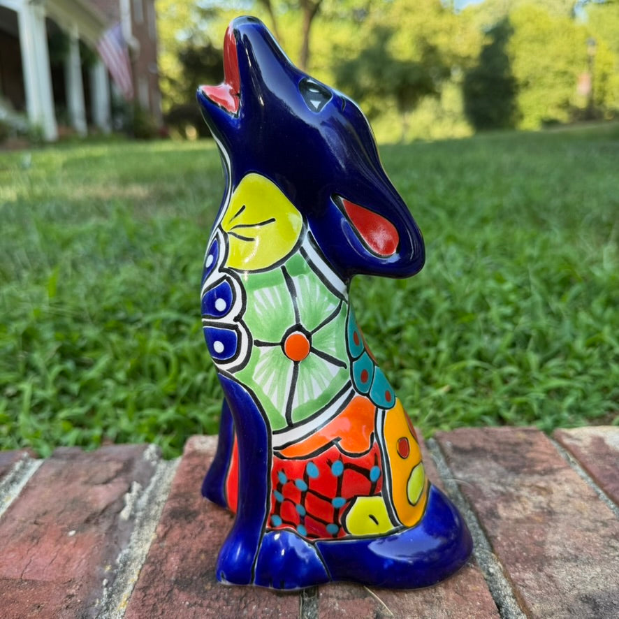 A colorful hand-painted talavera ceramic coyote figurine sitting on a brick surface with a blurred green lawn and trees in the background side view