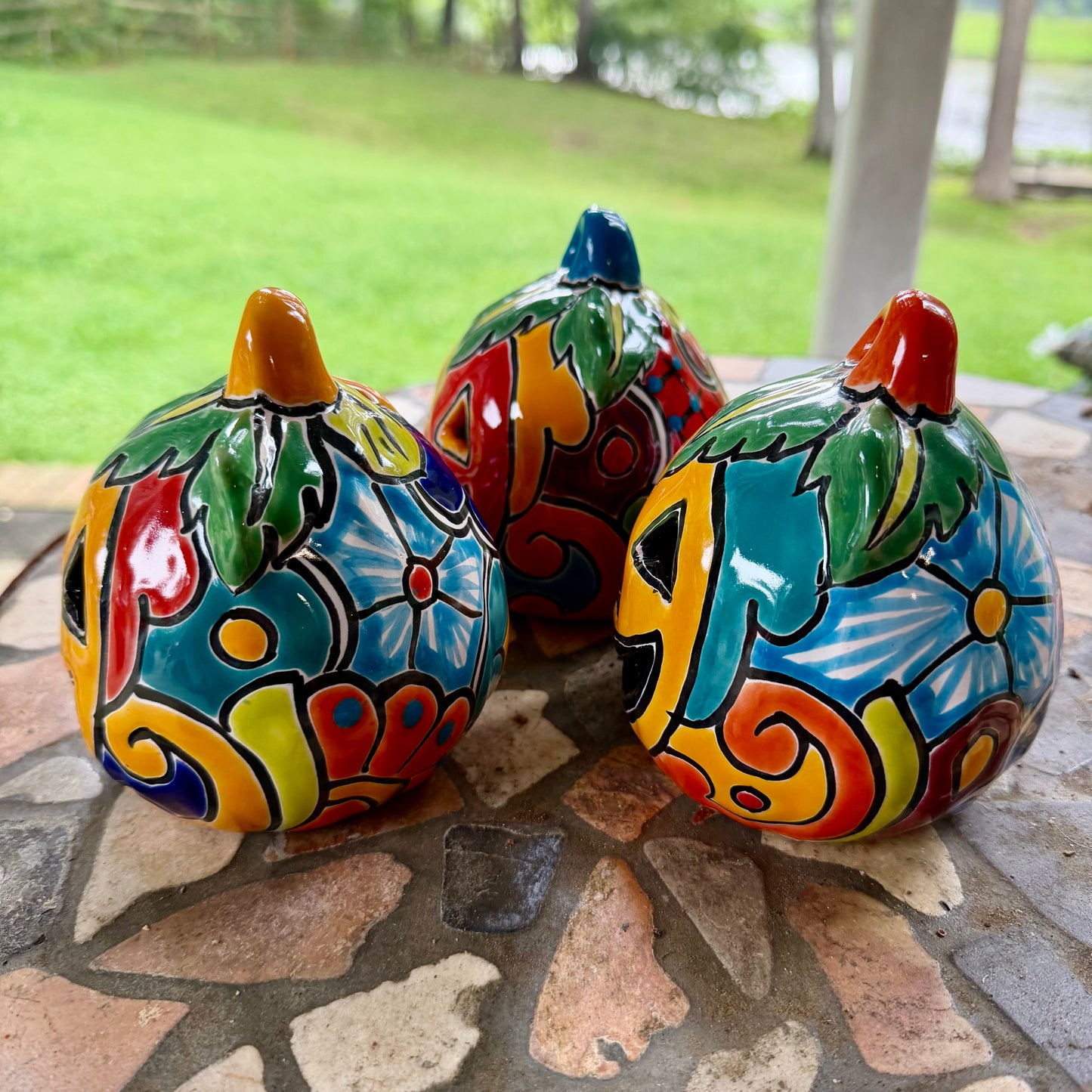 Three colorful hand-painted Talavera jack-o'-lantern pumpkins with multi-color displayed on a stone patio with a blurred green lawn background, left side view.