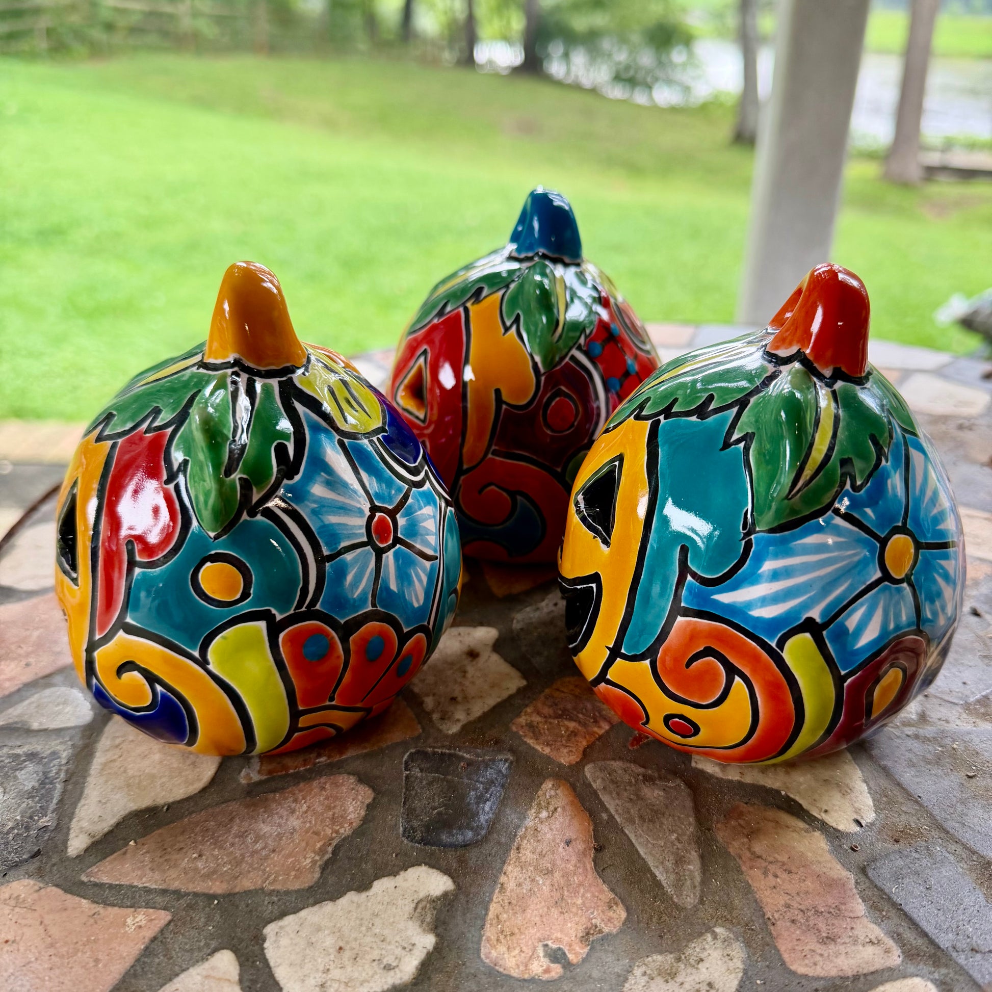 Three colorful hand-painted Talavera jack-o'-lantern pumpkins with multi-color displayed on a stone patio with a blurred green lawn background, left side view.