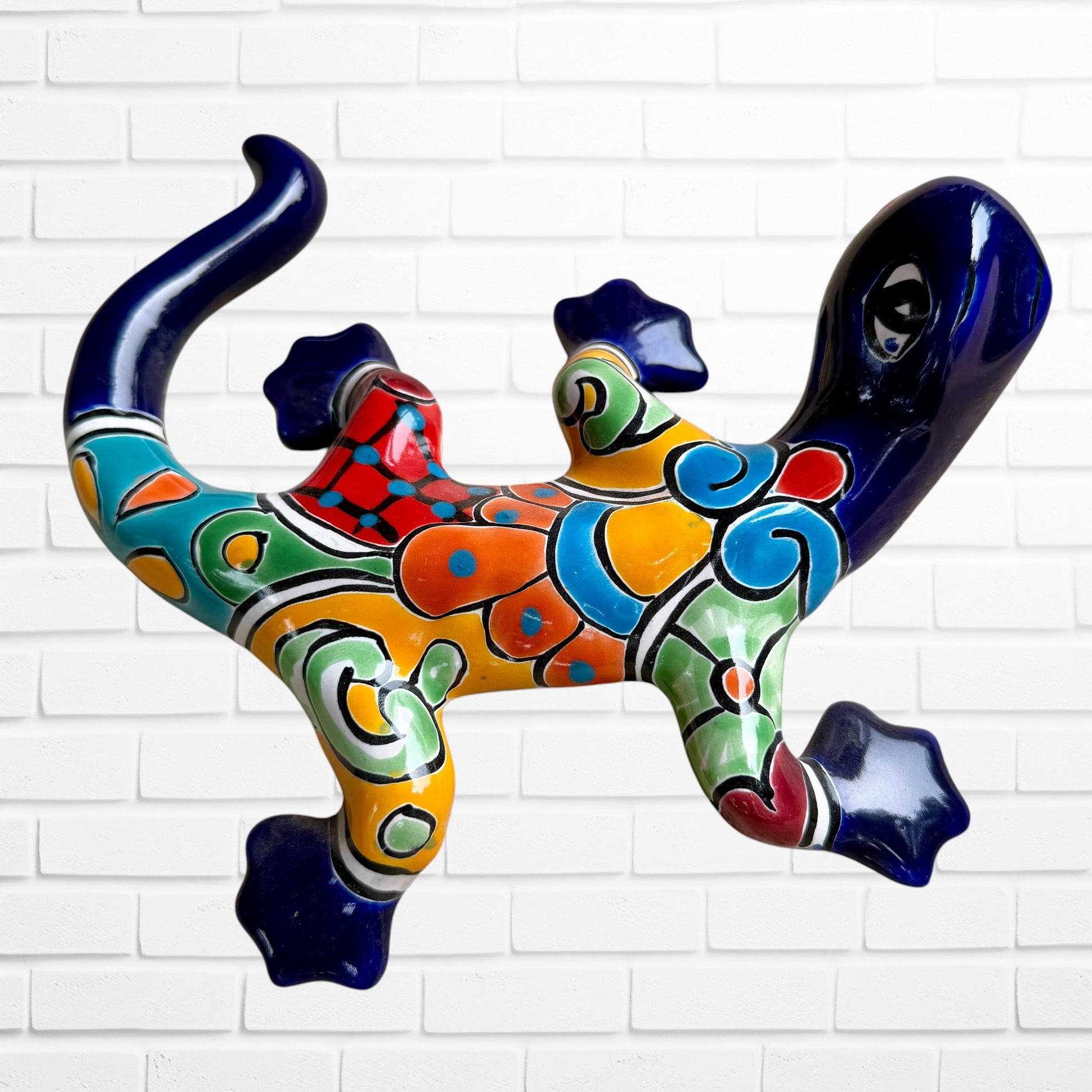 A colorful ceramic wall sculpture of a stylized lizard, decorated with vibrant hand-painted patterns in blue, orange, green, yellow, and red, mounted on a white brick wall back view