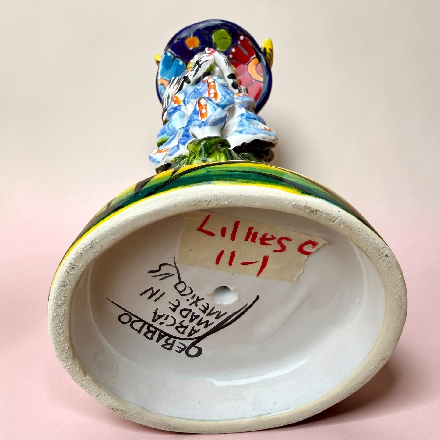 A Talavera Day of the Dead (Día de los Muertos) ceramic skeleton figurine with calla Lillies standing on a patterned base against a soft pink gradient background. Bottom view showing artist signature.