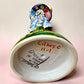 A Talavera Day of the Dead (Día de los Muertos) ceramic skeleton figurine with calla Lillies standing on a patterned base against a soft pink gradient background. Bottom view showing artist signature.