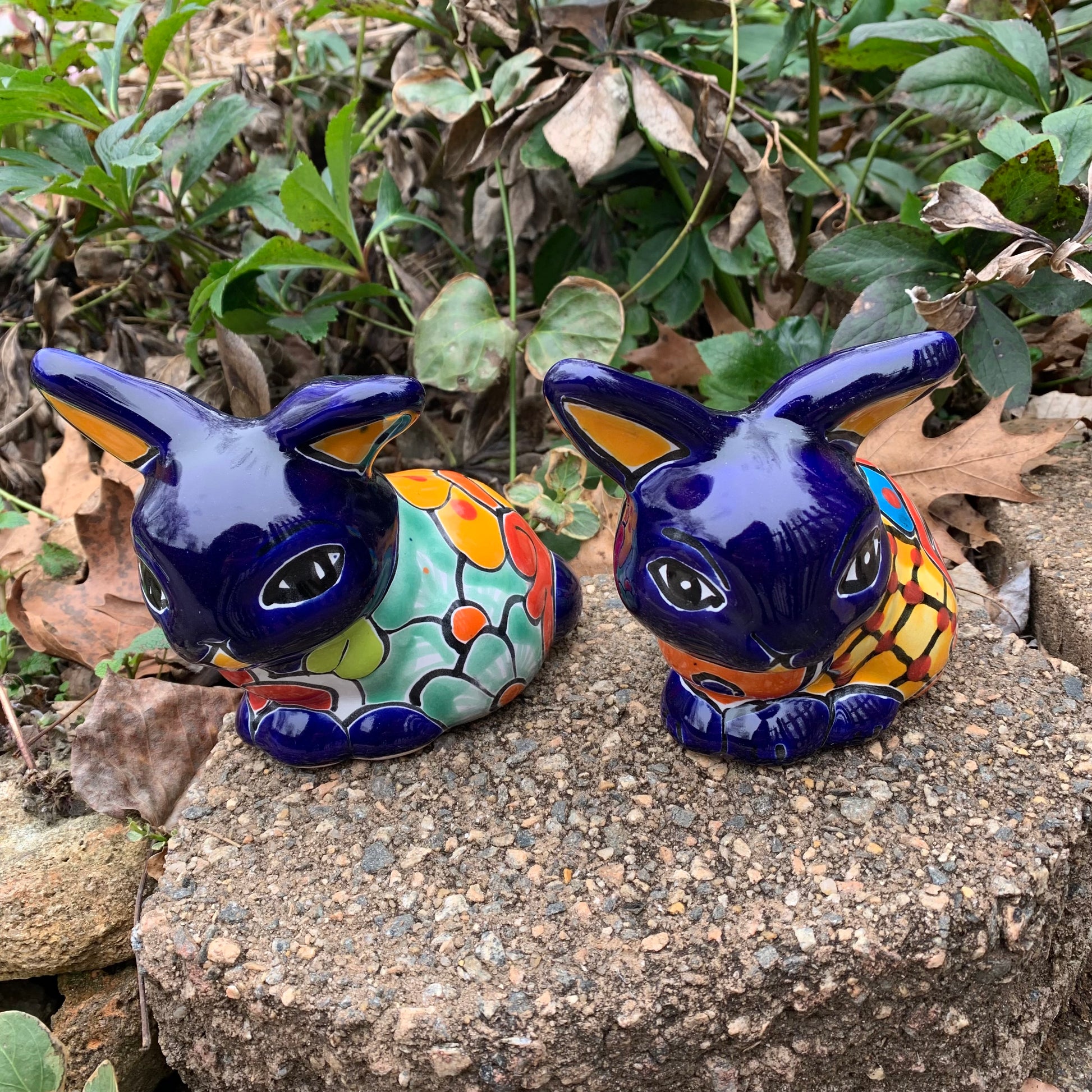 Two colorful Mexican Talavera rabbit figurines on a stone surface with greenery in the background in Azul front