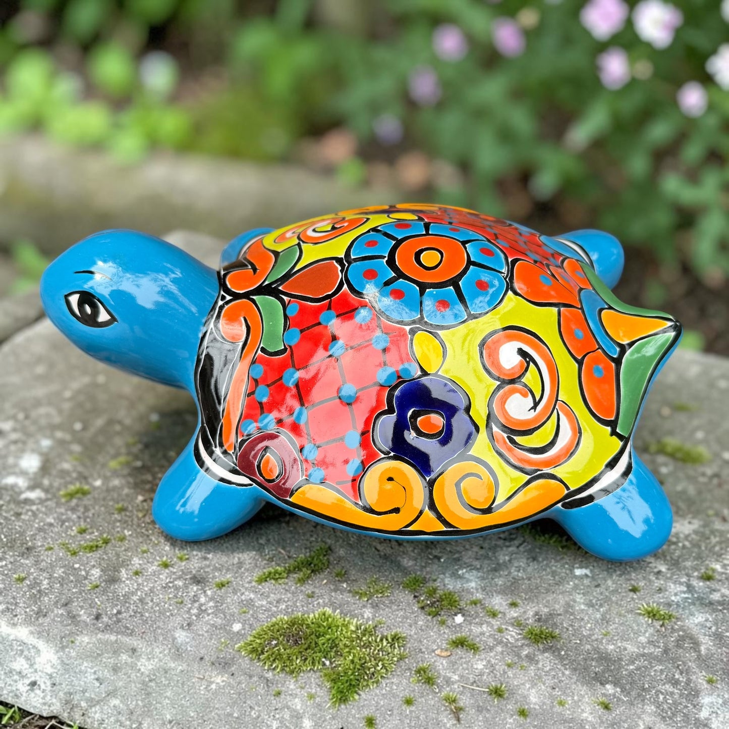 A hand-painted ceramic turtle figurine resting on a mossy stone outdoors, featuring a bright blue body and a highly decorated shell with vibrant orange, yellow, red, and blue patterns, right side view.