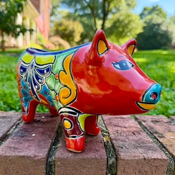 A colorful hand-painted red Talavera ceramic pig figurine standing on a brick surface with a blurred green lawn and trees in the background.