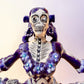 A Day of the Dead style talavera figurine of a skeleton woman in a wide floral skirt, painted in deep blue, black, white, and light blue on a soft beige-to-blue gradient background. Close up of ballerina face.