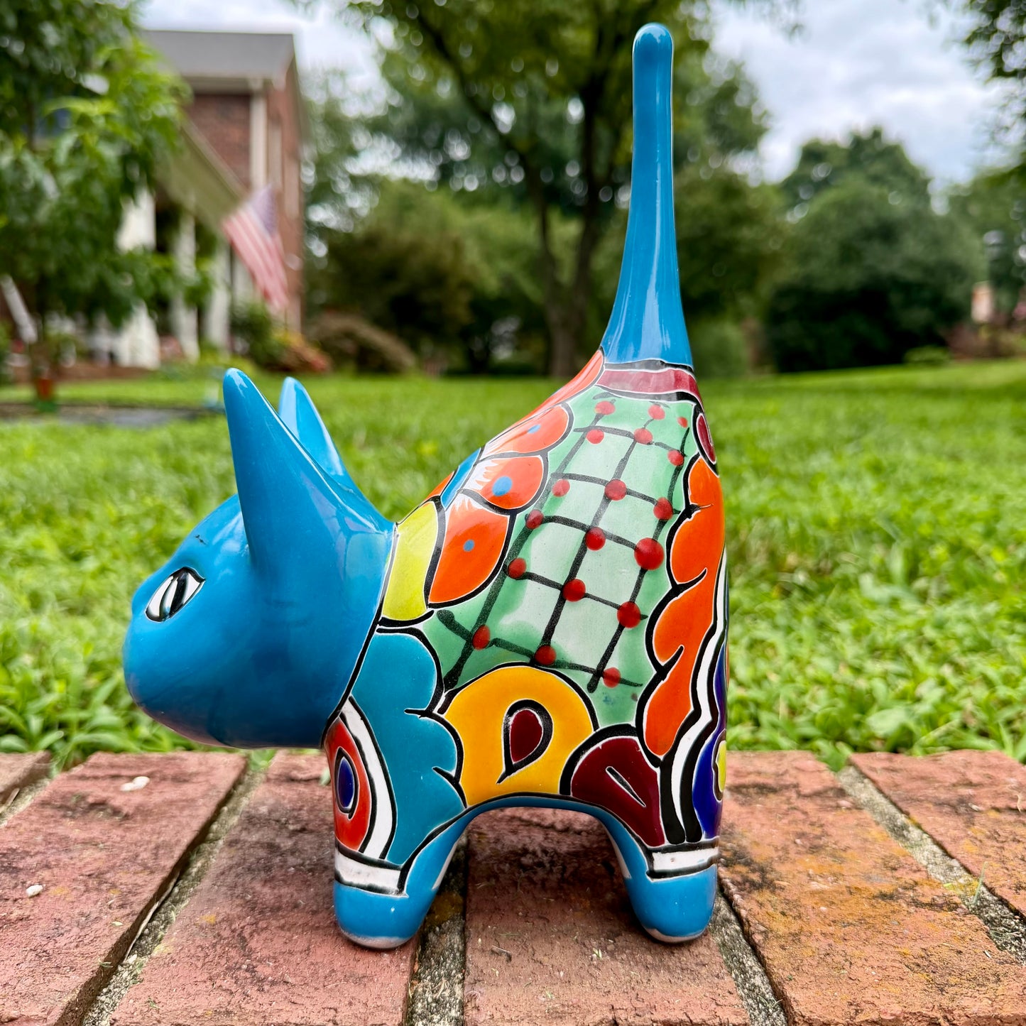 A glossy talavera ceramic cat figurine decorated with vibrant hand-painted patterns in turquoise, yellow, green and red, standing on a brick path with a green lawn and a house in the blurred background right side view.