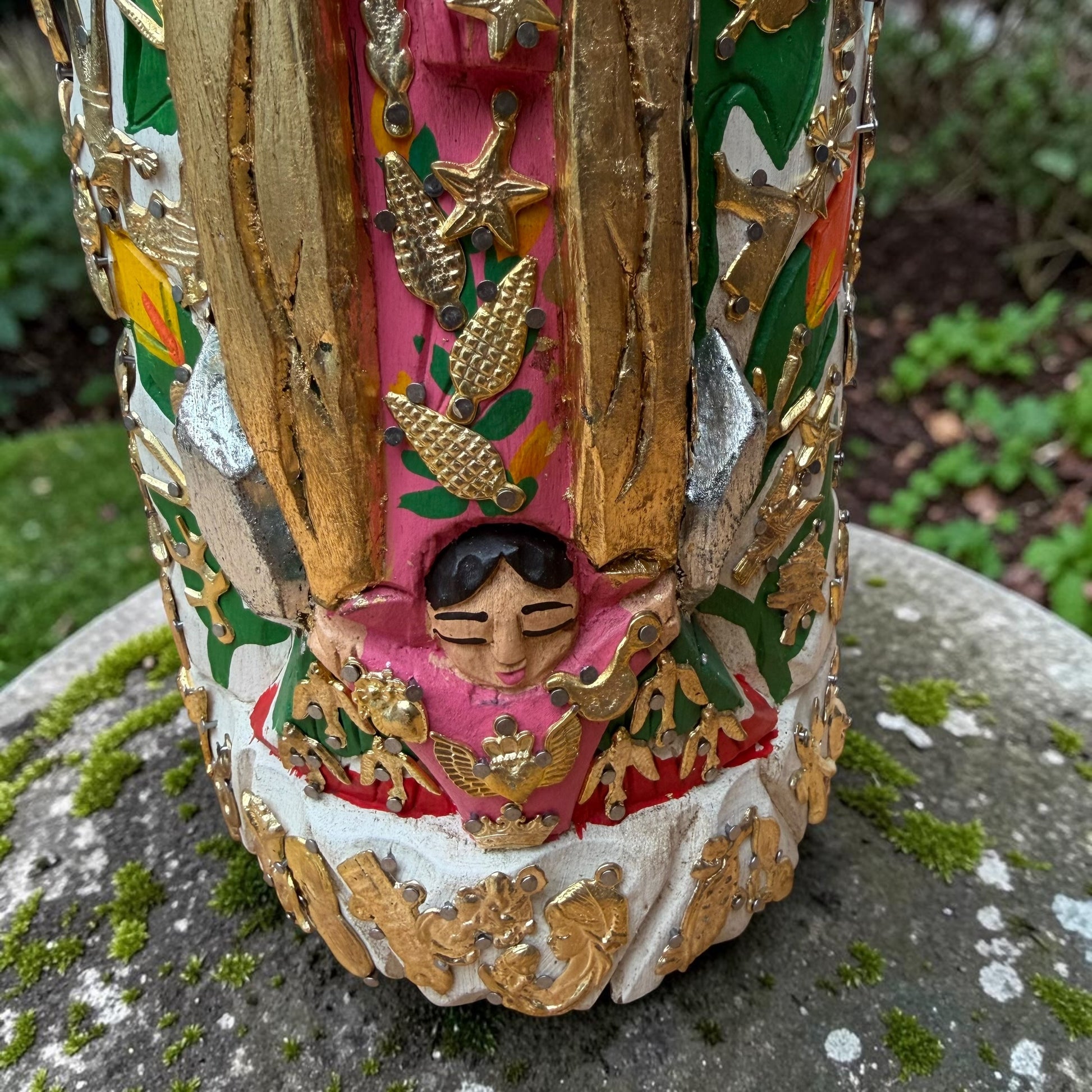 A Milagros religious figurine of Our Lady of Guadalupe with a child, standing on a mossy stone surface with a blurred outdoor garden background. She has a white halo and Lillies adorning the halo. Child Close up.