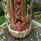 A Milagros religious figurine of Our Lady of Guadalupe with a child, standing on a mossy stone surface with a blurred outdoor garden background. She has a white halo and Lillies adorning the halo. Child Close up.