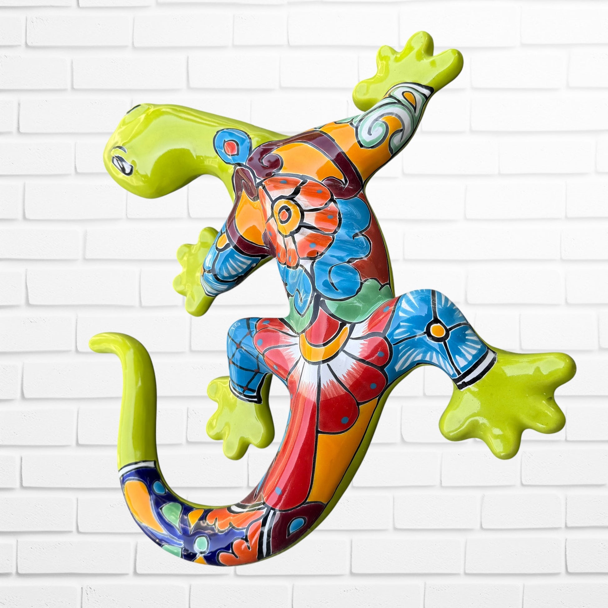 A colorful decorative lizard Talavera wall sculpture with a hand-painted floral and geometric pattern, mounted against a white brick wall, right side view.