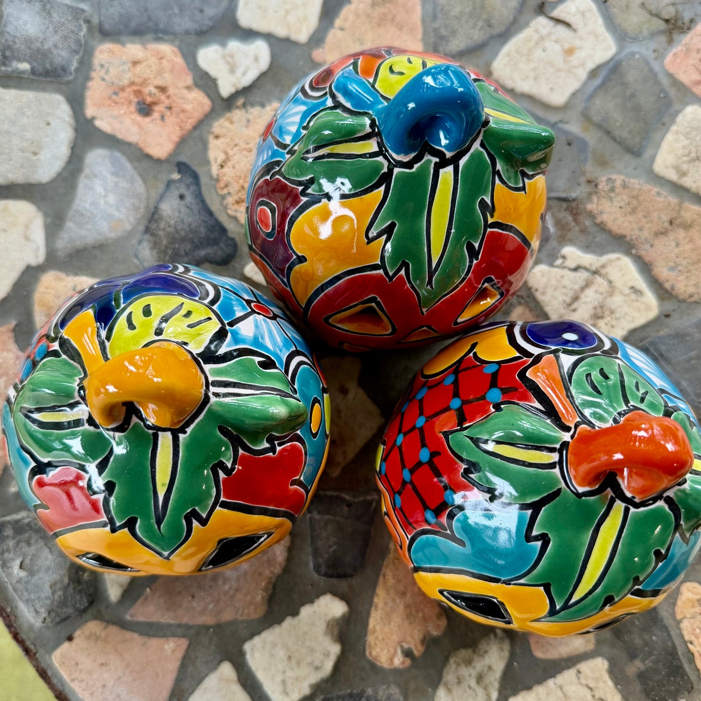 Three colorful hand-painted Talavera jack-o'-lantern pumpkins with multi-color displayed on a stone patio with a blurred green lawn background, top stem view.