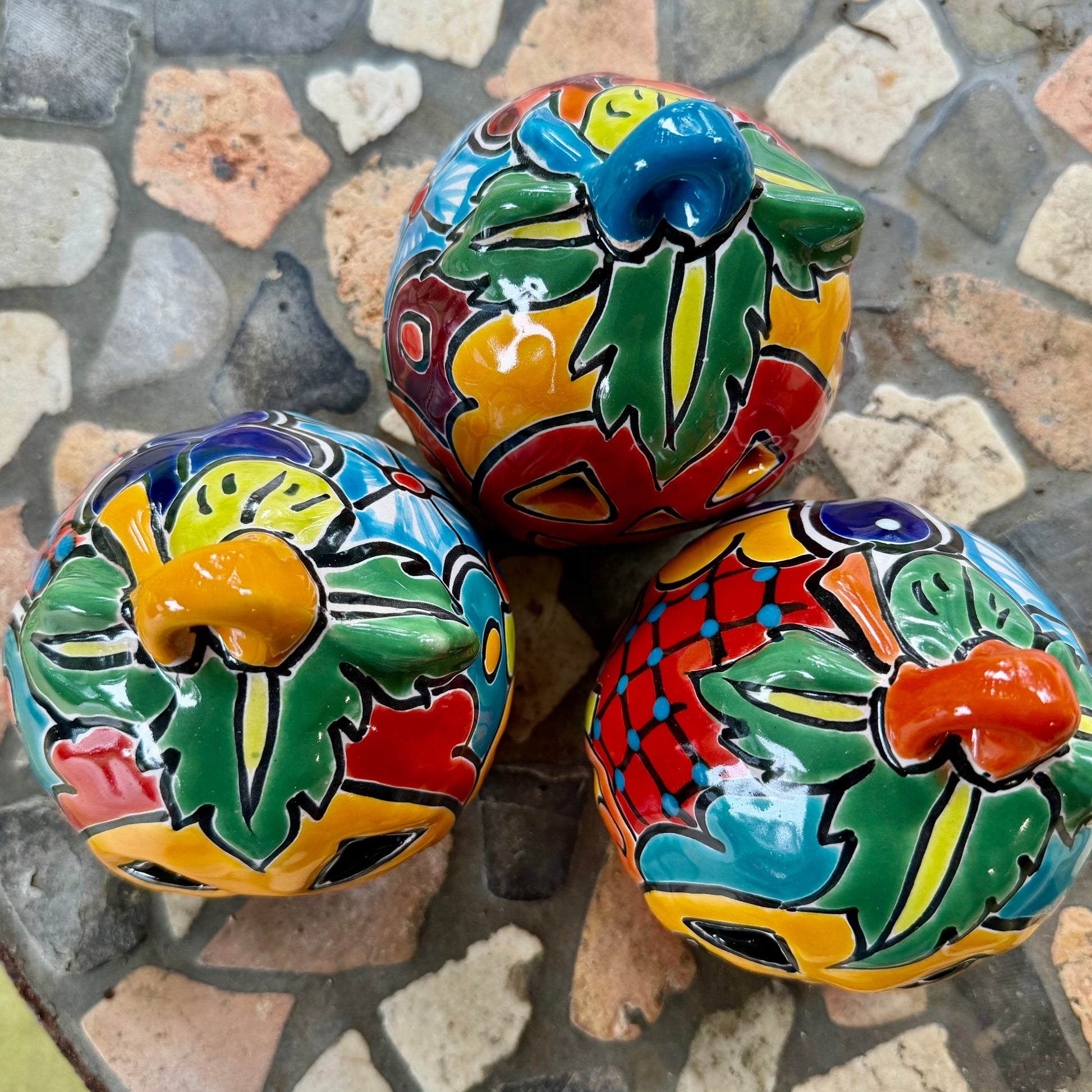 Three colorful hand-painted Talavera jack-o'-lantern pumpkins with multi-color displayed on a stone patio with a blurred green lawn background, top stem view.