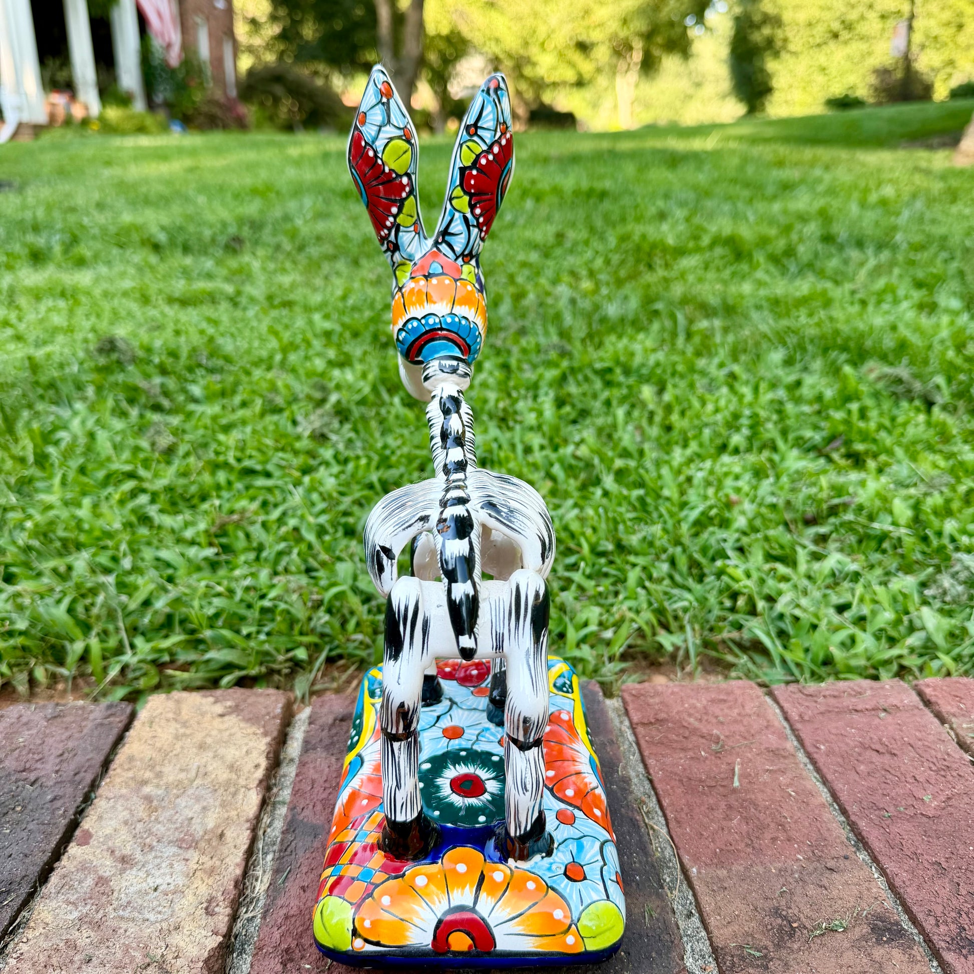 A colorful hand-painted Talavera Catrina donkey figurine, standing on a colorful floral base in an outdoor backyard setting, view of the back tail.