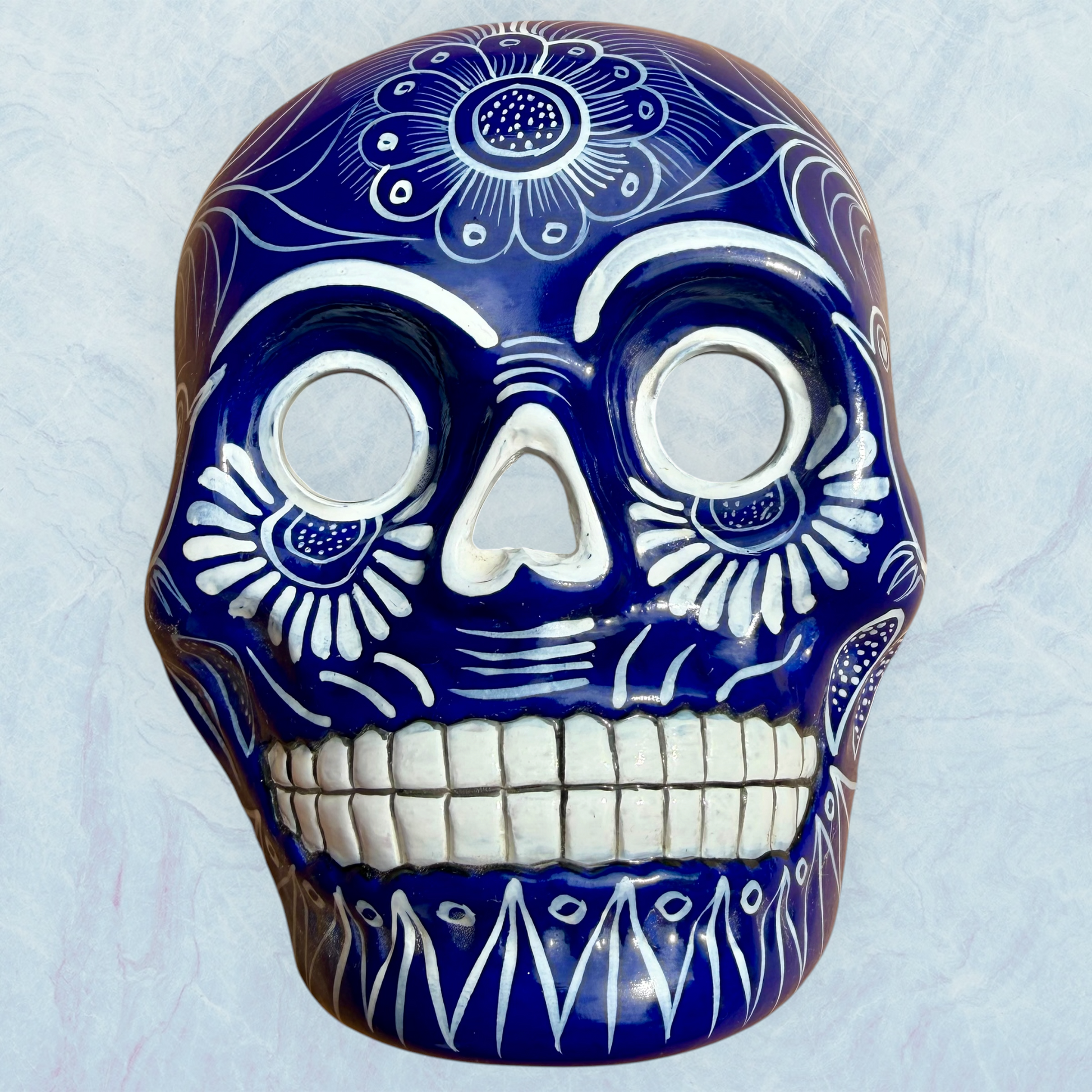 A clay, blue with white designs, Mexican-style skeleton skull face mask hanging on a soft blue surface.