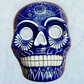 A clay, blue with white designs, Mexican-style skeleton skull face mask hanging on a soft blue surface.