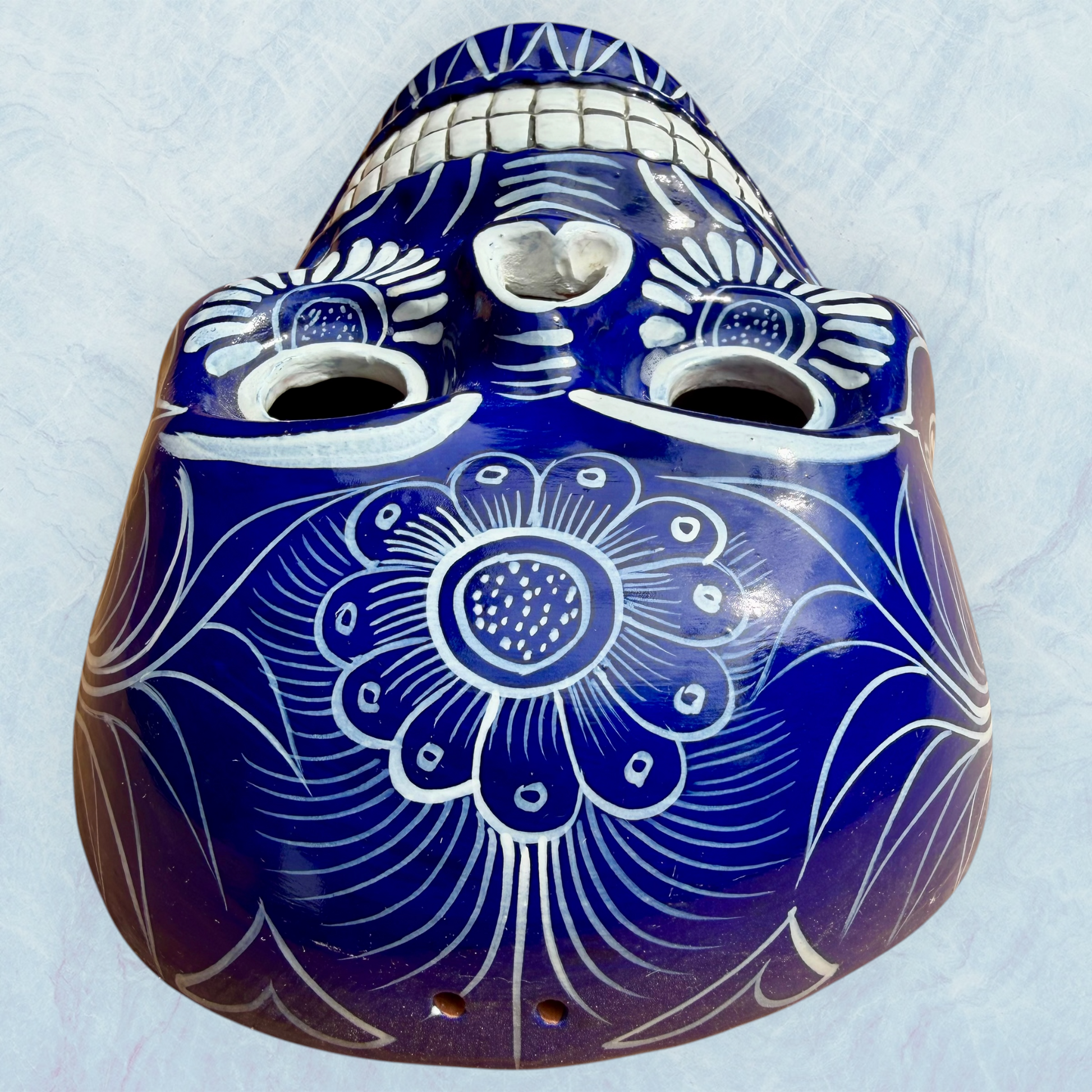 A clay, blue with white designs, Mexican-style skeleton skull face mask hanging on a soft blue surface, top forehead view.