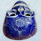 A clay, blue with white designs, Mexican-style skeleton skull face mask hanging on a soft blue surface, top forehead view.