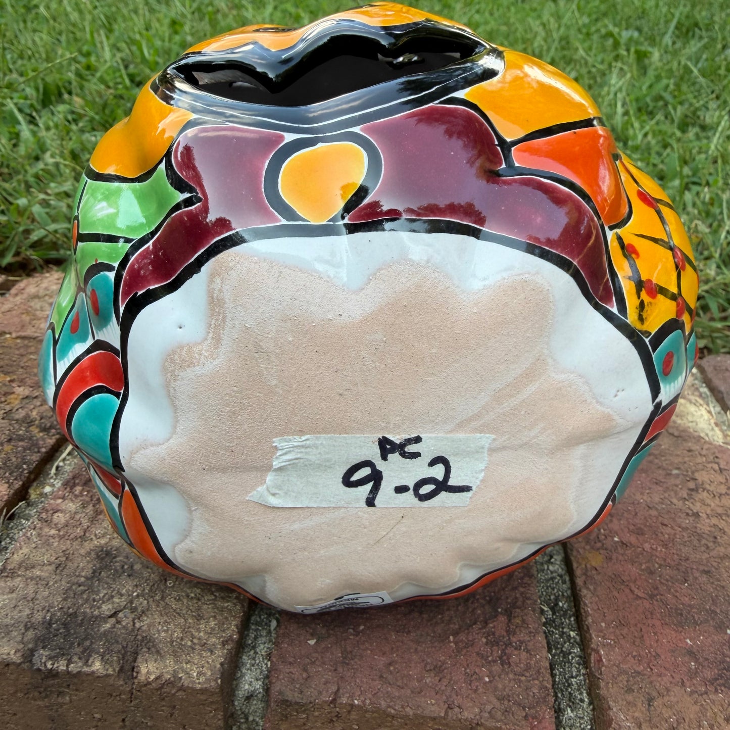 A Talavera jack-o'-lantern pumpkin decoration with a carved face, sitting on a brick surface in an outdoor yard with grass and trees in the background. Bottom of the pumpkin.
