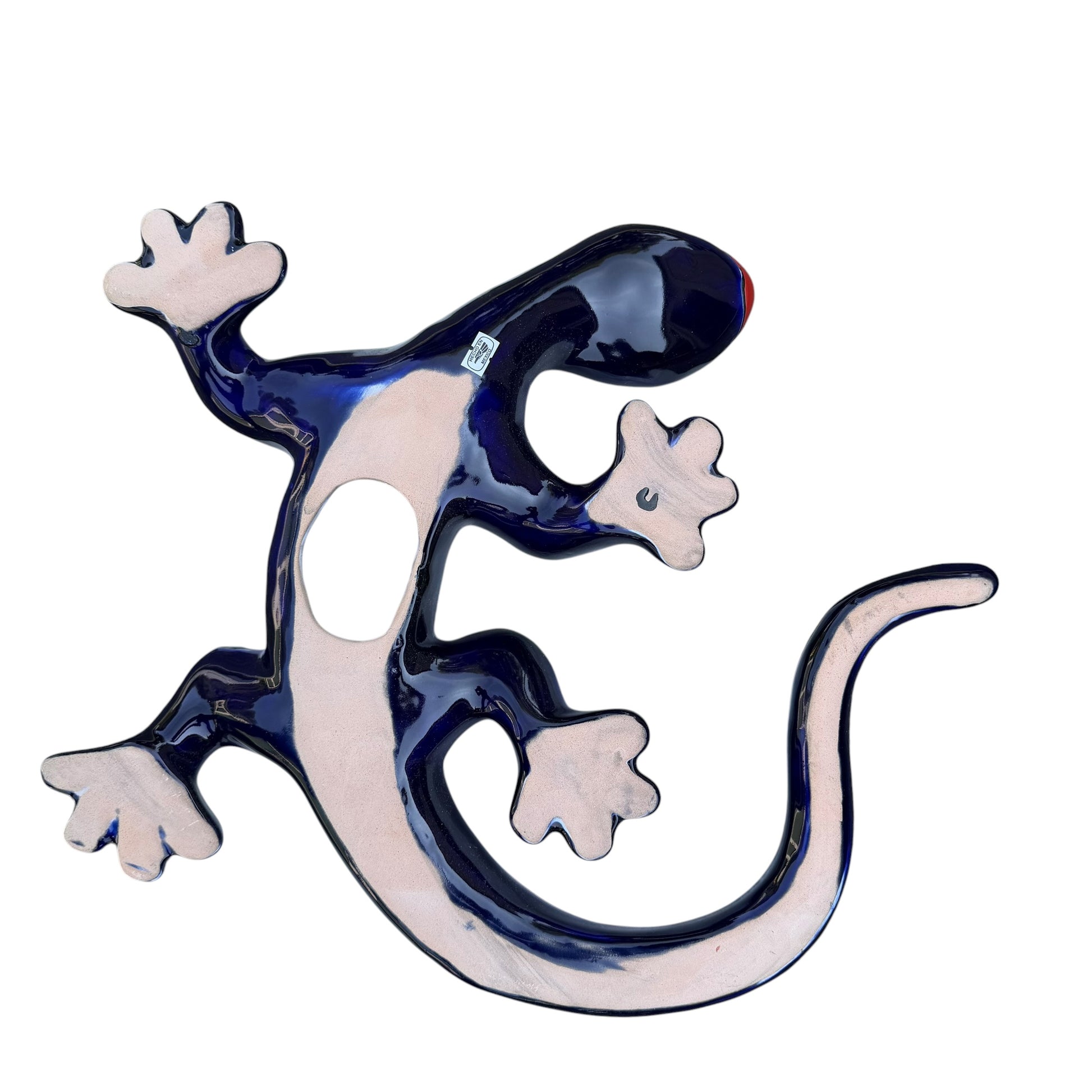 A colorful Mexican Talavera wall sculpture of a lizard, featuring a vibrant multicolor pattern with dark blue, orange, red, green, and yellow, mounted on a white brick wall, back showing hanging hole.