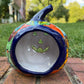 A Talavera jack-o'-lantern pumpkin decoration with a carved face, sitting on a brick surface in an outdoor yard with grass and trees in the background. Parkview Showing opening for candle.