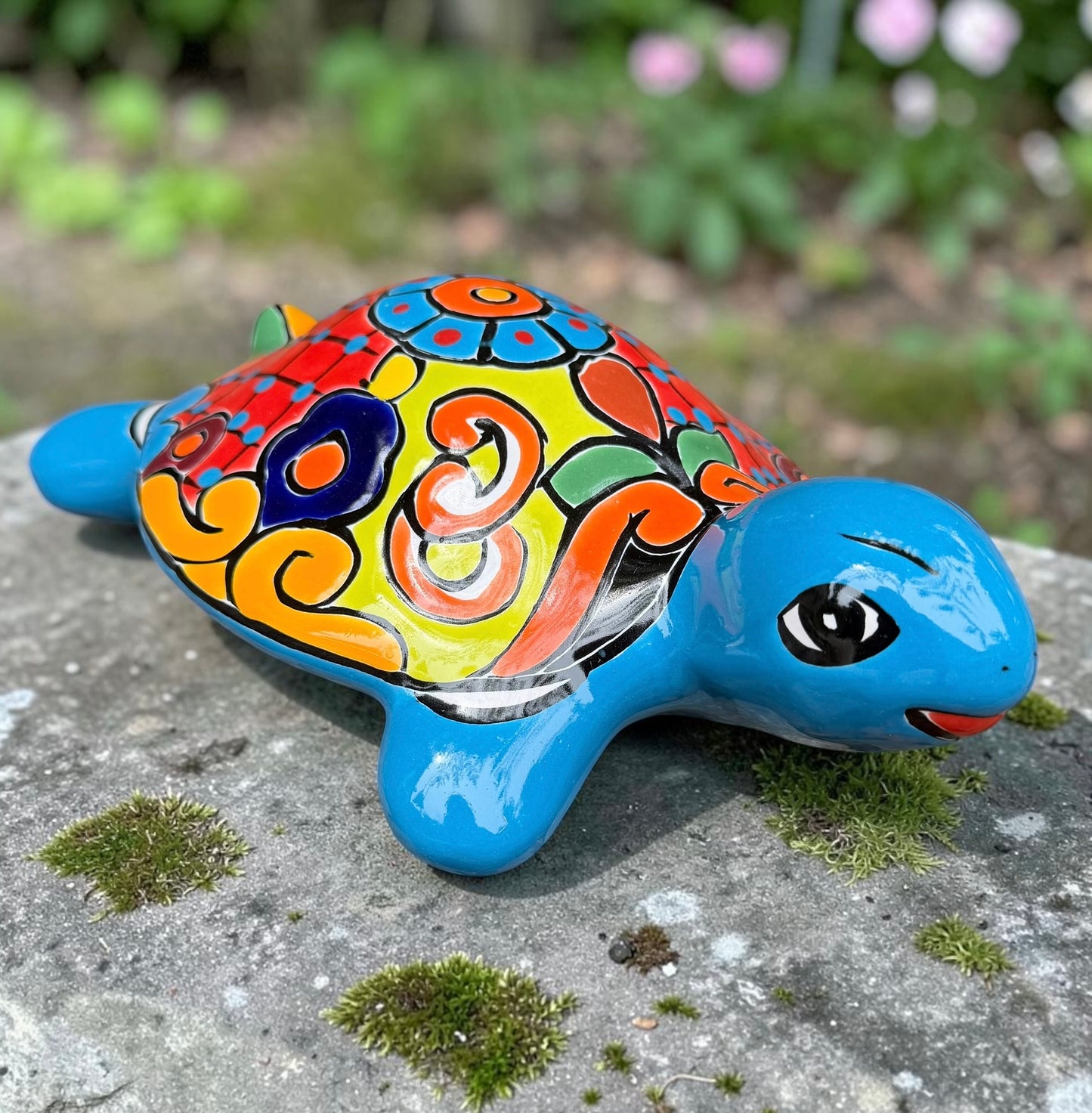 A hand-painted ceramic turtle figurine resting on a mossy stone outdoors, featuring a bright blue body and a highly decorated shell with vibrant orange, yellow, red, and blue patterns.