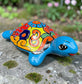 A hand-painted ceramic turtle figurine resting on a mossy stone outdoors, featuring a bright blue body and a highly decorated shell with vibrant orange, yellow, red, and blue patterns.