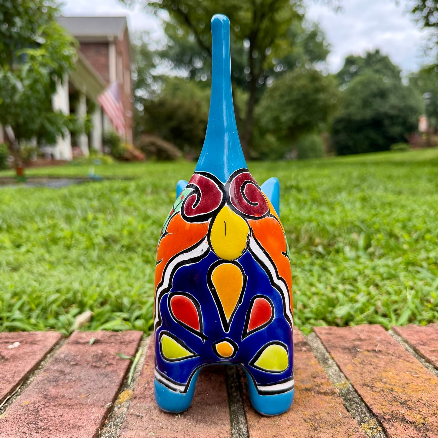 A glossy talavera ceramic cat figurine decorated with vibrant hand-painted patterns in turquoise, yellow, green and red, standing on a brick path with a green lawn and a house in the blurred background. Back tail view.