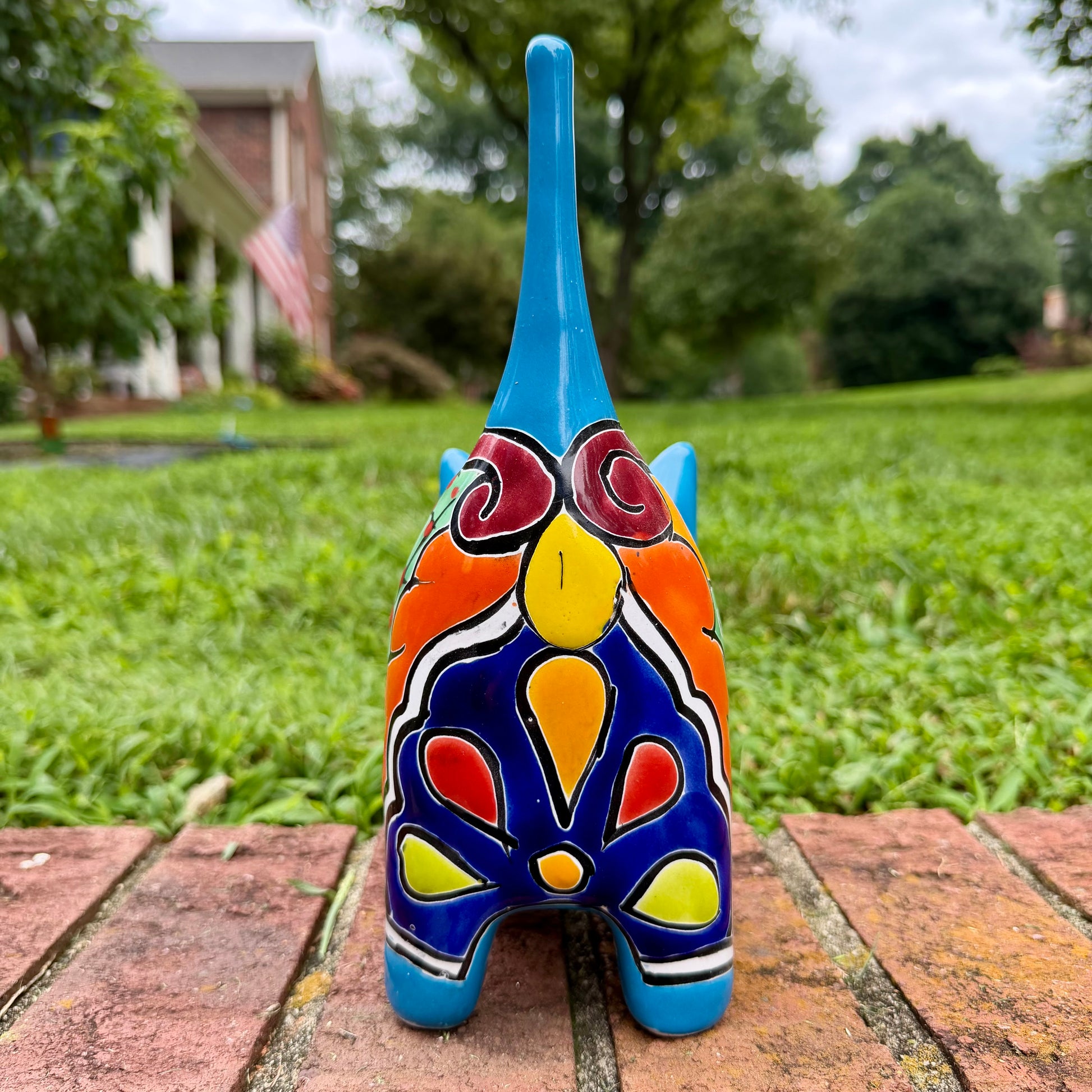 A glossy talavera ceramic cat figurine decorated with vibrant hand-painted patterns in turquoise, yellow, green and red, standing on a brick path with a green lawn and a house in the blurred background. Back tail view.